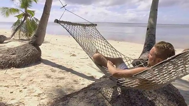 年轻男子在菲律宾热带岛屿海滩的吊床上放松的慢动作视频。人们旅游度假放松的概念。