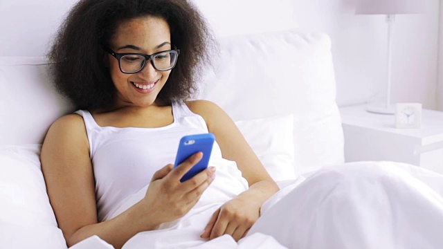 年轻的非洲妇女在家里卧室的床上用智能手机发短信