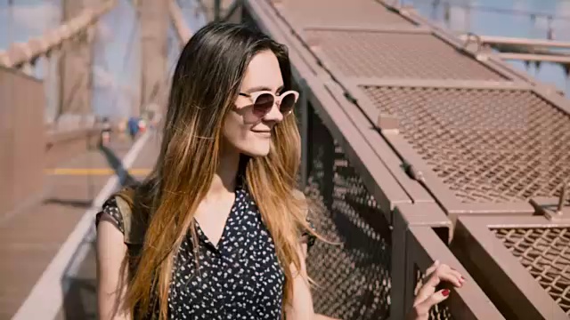 详情图-美丽的欧洲女人戴着时尚的太阳镜走向相机，触摸头发在布鲁克林大桥的影子护栏4K