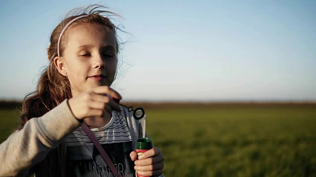在一个阳光明媚的日子里，可爱的小女孩正在草地上吹肥皂泡。缓慢的运动。快乐的童年。模糊的背景
