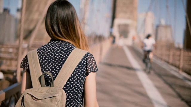 镜头跟随女游客背包和深色金发沿着布鲁克林大桥走在一个晴朗的晴天4K