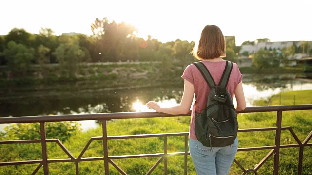 年轻迷人的女孩背着背包站在桥上，看着夕阳下的河水