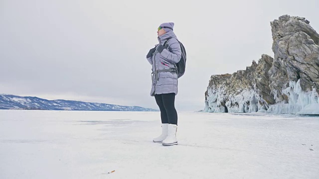 女人在贝加尔湖冰面上的旅行。冬岛之旅。女孩在冰岩石下行走。游客看着美丽的冰洞。极限跋涉和步行。背包客在大自然中休憩。