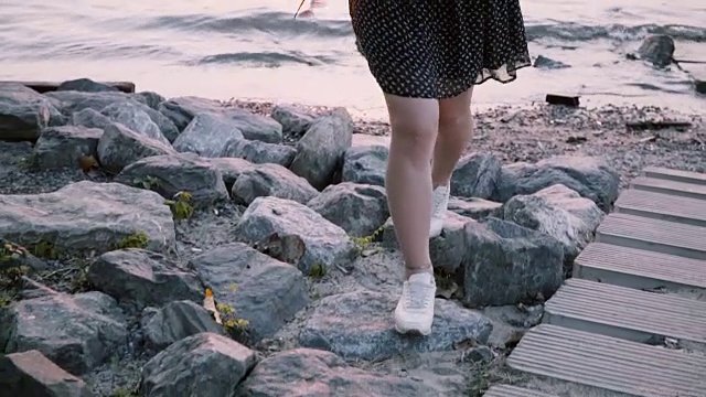 美丽的年轻女孩穿着黑色的衣服走在大河边的石头上，在夏季城市的夕阳下，倾斜下来的慢动作