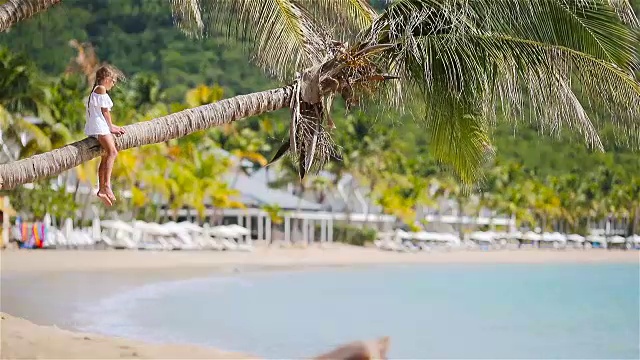 可爱的小女孩坐在棕榈树上在白色的海滩暑假