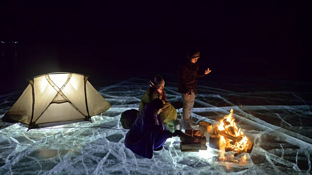 三个旅行者在夜间的冰面上着火。营地在冰上。帐篷紧挨着火。贝加尔湖。附近有一辆车。人们围着篝火取暖，穿着睡袋。
