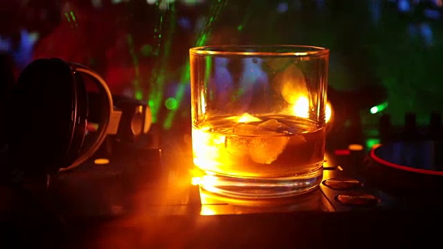 在夜总会的dj控制器上，装有冰块的威士忌酒杯。在迪斯科灯光下的夜总会音乐派对上，一杯威士忌上戴着Dj耳机。滑块拍摄