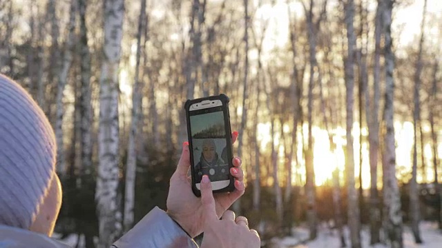 一个女人走在树林里。旅行者在森林里用手机拍照。女孩会自拍，用智能手机交流。美丽多汁的日落时分。
