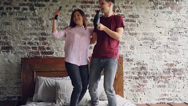 詳情圖-好玩的年輕人男人和女人在床上跳舞聽音樂和唱歌用吹風(fēng)機和電視遙控器作為麥克風(fēng)一起玩。