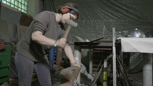 女铁匠戴着保护耳机，在她的diy车间里用锤子敲打金属棒来把它弄直