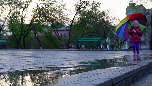 详情图-可爱的小女孩拿着五颜六色的雨伞在夕阳下穿过公园的水坑