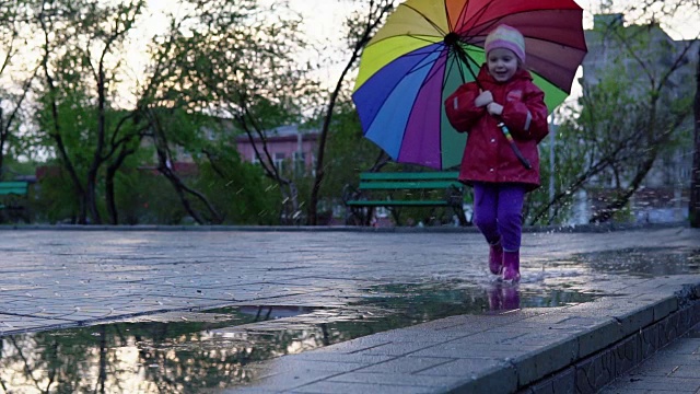 详情图-可爱的小女孩拿着五颜六色的雨伞在夕阳下穿过公园的水坑