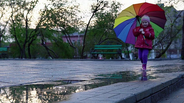详情图-可爱的小女孩拿着五颜六色的雨伞在夕阳下穿过公园的水坑