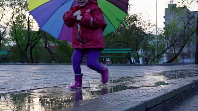 详情图-可爱的小女孩拿着五颜六色的雨伞在夕阳下穿过公园的水坑