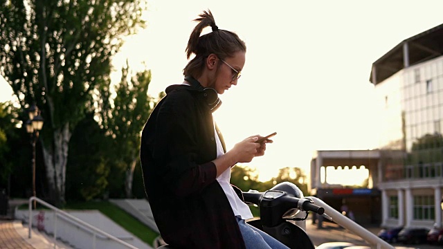 穿着黑色连帽衫的年轻女孩坐在摩托车上用手机打字。坐在公园外面的摩托车上。阳光照射在背景上