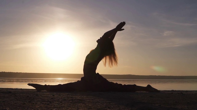 日出时海滩上一个美丽的瑜伽女孩的剪影。Hanumanasana,猴子姿势。阳光洒在一具身体上