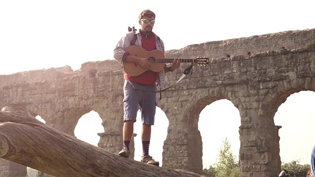 年轻的男子冒险家旅行者站在一个原木树干上弹吉他唱歌在古罗马渡槽遗址在parco degli acquedotti公园在罗马日出慢镜头