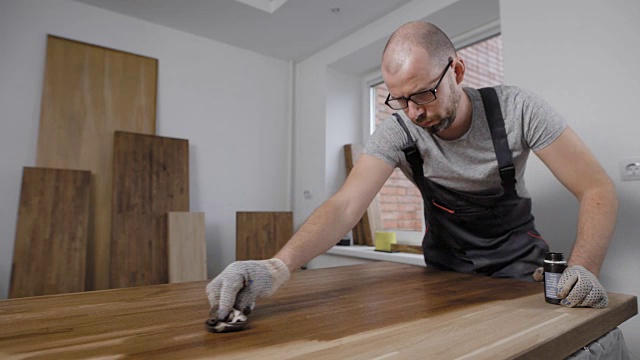 一名成年男子正在他家里的一个房间里用清漆覆盖木板，做木工和组装家具