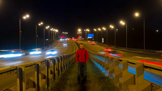 一个人站在公路上。随着时间的推移，高速公路的交通使汽车在夜间变得模糊不清。汽车在桥上行驶的晚上时间流逝。4 k UHD