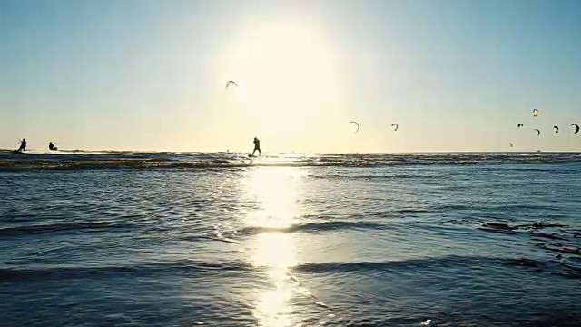 风筝冲浪在夏天的海上在有风的天气日落。摄像机沿着海岸移动，慢镜头