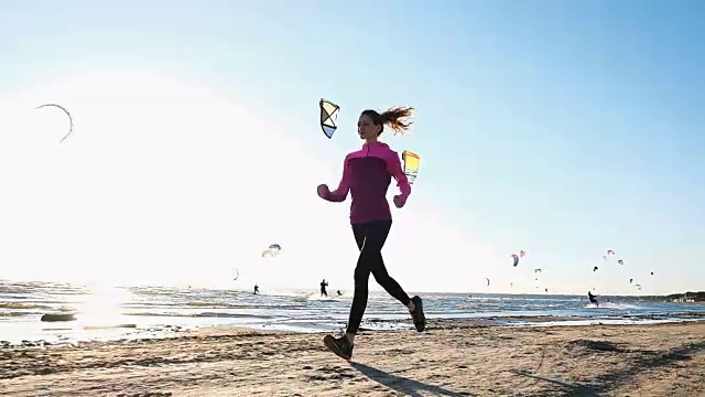 年轻女子在夏天的海滩上跑步，背景是风筝冲浪者，慢动作