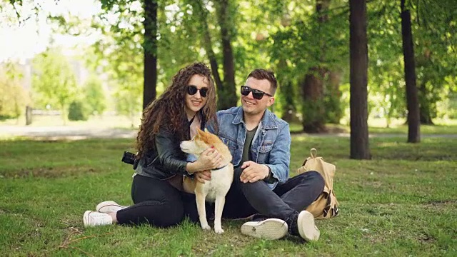 兴奋的年轻人恩爱夫妇正在逗可爱的日本柴犬挠它的毛，在公园的草地上有说有笑。人与动物的概念。