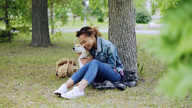 活泼的混血女孩拥抱着美丽的日本柴犬，抚摸着在公园树下休息的动物，而宠物享受着爱和照顾。