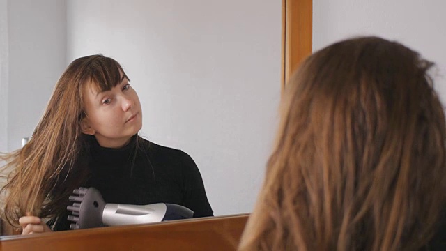 年轻迷人的棕发女子微笑着在家里镜子前用吹风机吹干她飘动的头发。