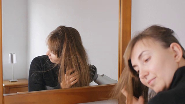 年轻迷人的棕发女子微笑着在家里的镜子前用吹风机吹干她的头发。