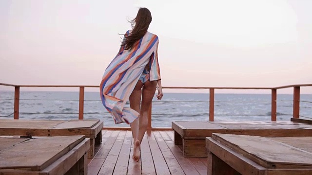 详情图-华丽的背后慢动作镜头，一个白人模特在泳衣和夏季开襟羊毛衫走在户外的木地板上，风摇动她的布料。大海或海洋的背景